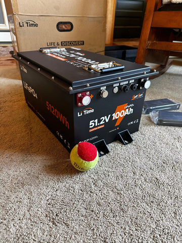 A LiTime 51.2V 100Ah battery is placed on carpet, showcasing its features alongside a tennis ball for scale, in a customer review
