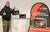 A man gives a thumbs-up while holding a LiTime 12.8V 100Ah battery in front of a promotional banner at an expo booth