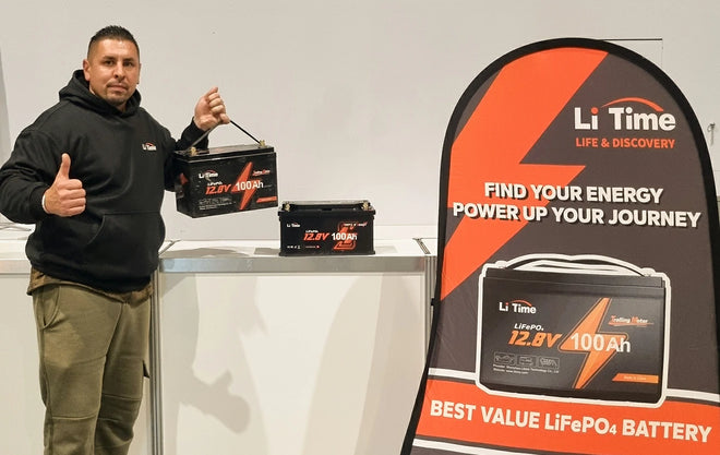 A man gives a thumbs-up while holding a LiTime 12.8V 100Ah battery in front of a promotional banner at an expo booth