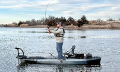 A man wearing a life vest stands in a fishing kayak, casting his line into the water