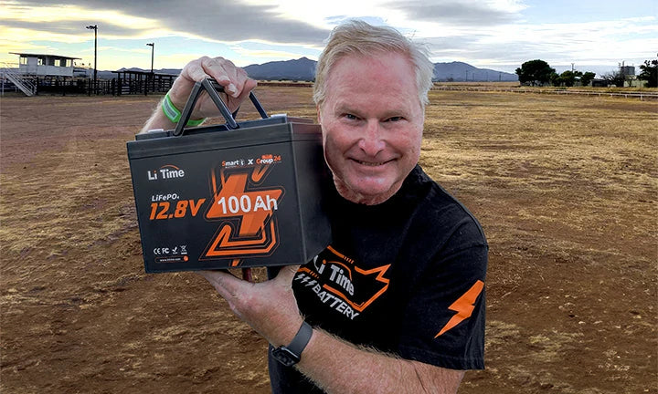 A man holds a LiTime 12.8V 100Ah battery outdoors, showcasing its features against a scenic desert backdrop