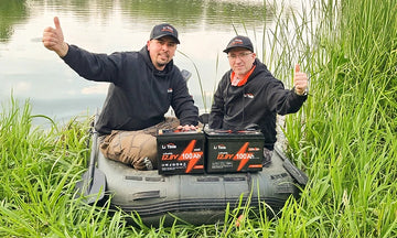 Two men give a thumbs-up while sitting in an inflatable boat on a grassy bank with two LiTime batteries